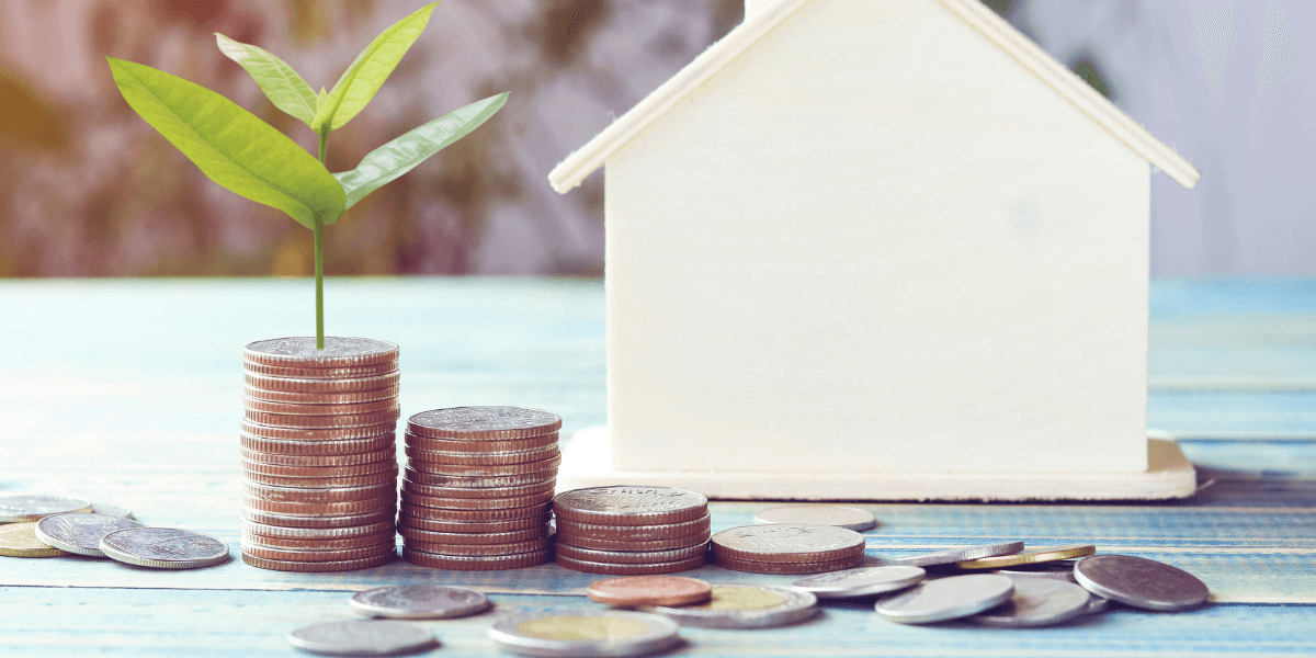 Stacks of coins with a plant growing on top beside a wooden house model