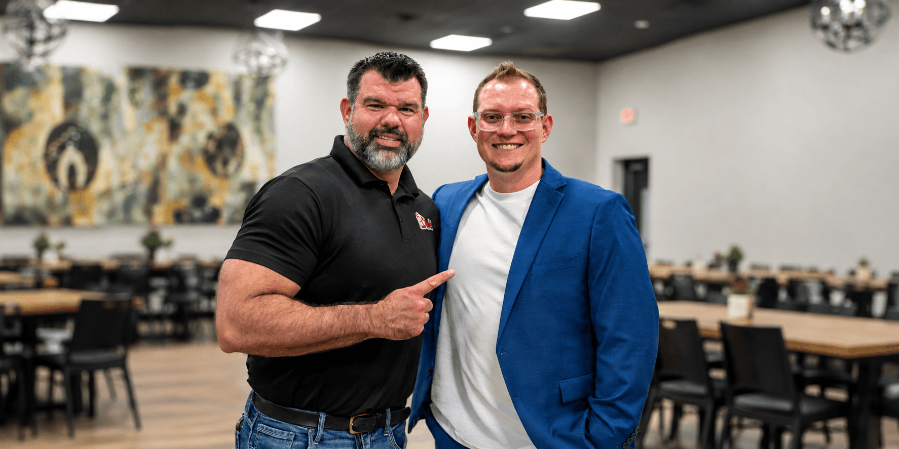Two men posing together in a restaurant setting, one in a black shirt and the other in a blue blazer.