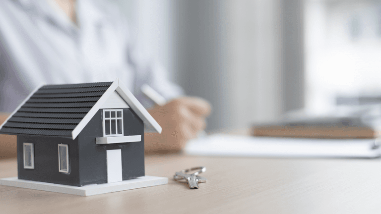 Model house and keys on a desk with a person reviewing real estate documents in the background.