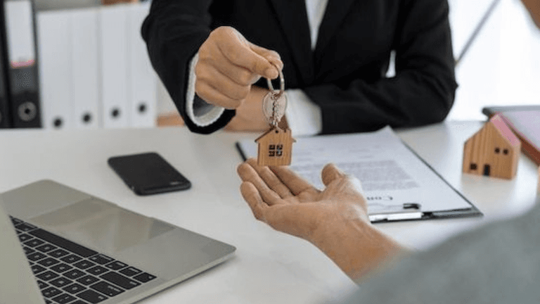 Real estate professional hands house keys to a buyer across a desk with miniature homes and a laptop.