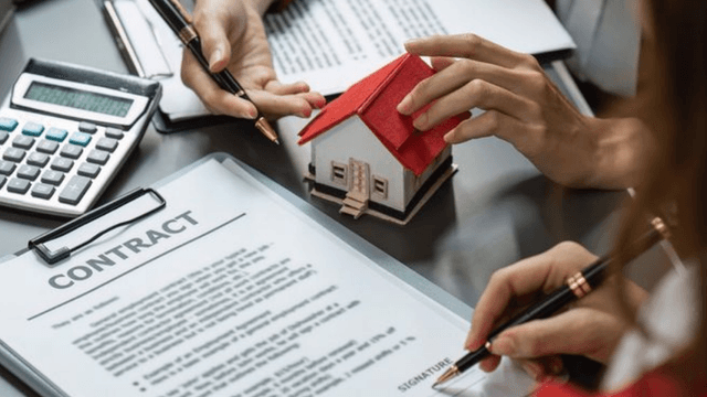 Hands holding a model house and pens over a real estate contract and calculator
