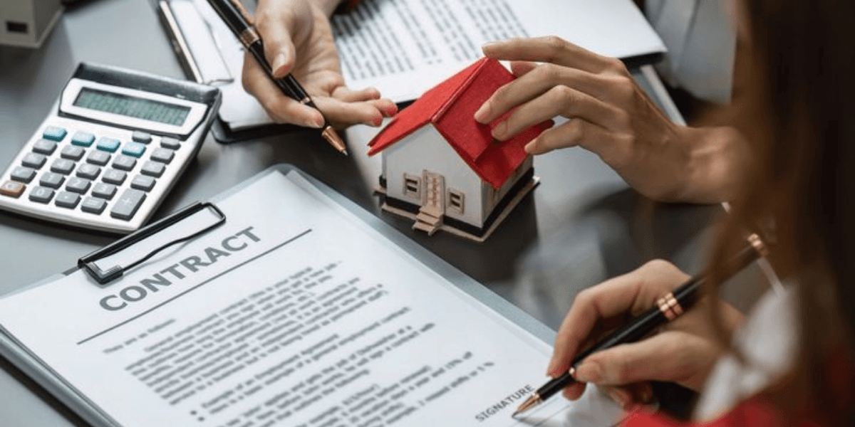 Hands holding a model house and pens over a real estate contract and calculator
