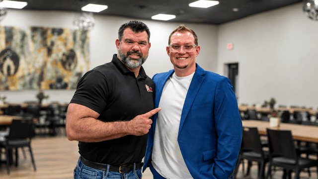 Two men posing together in a restaurant setting, one in a black shirt and the other in a blue blazer.