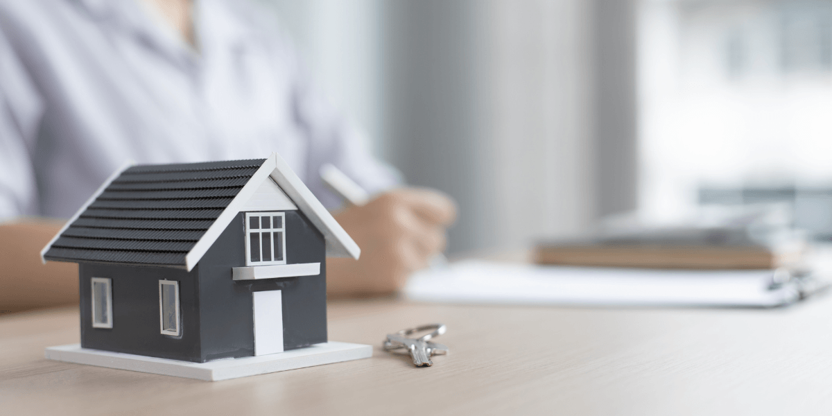 Model house and keys on a desk with a person reviewing real estate documents in the background.