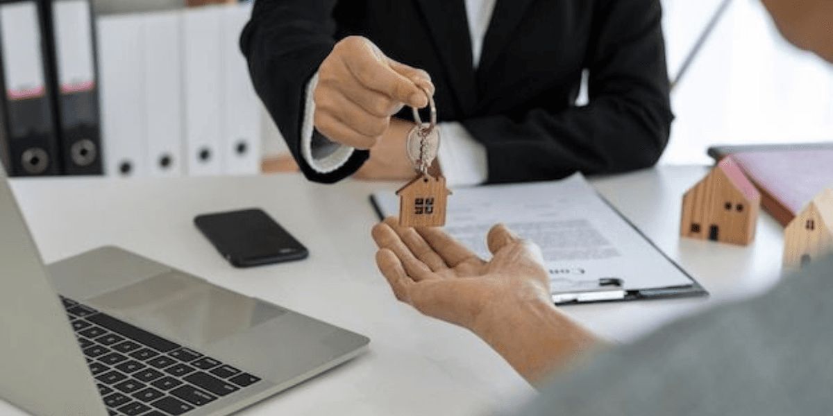Real estate professional hands house keys to a buyer across a desk with miniature homes and a laptop.